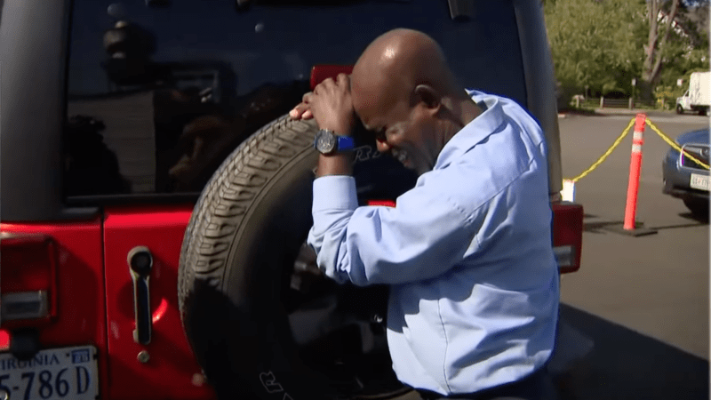 A man weeps in joy as he leans against the spare tire on the back of a red Jeep Wrangler that his students bought for him